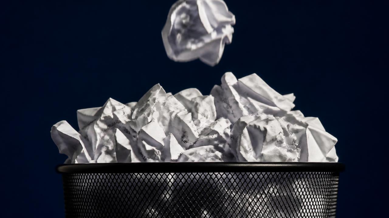 Wastebasket overflowing with crumpled paper drafts and one more falling in, illustrating that critique is care and iteration happens before work ships.